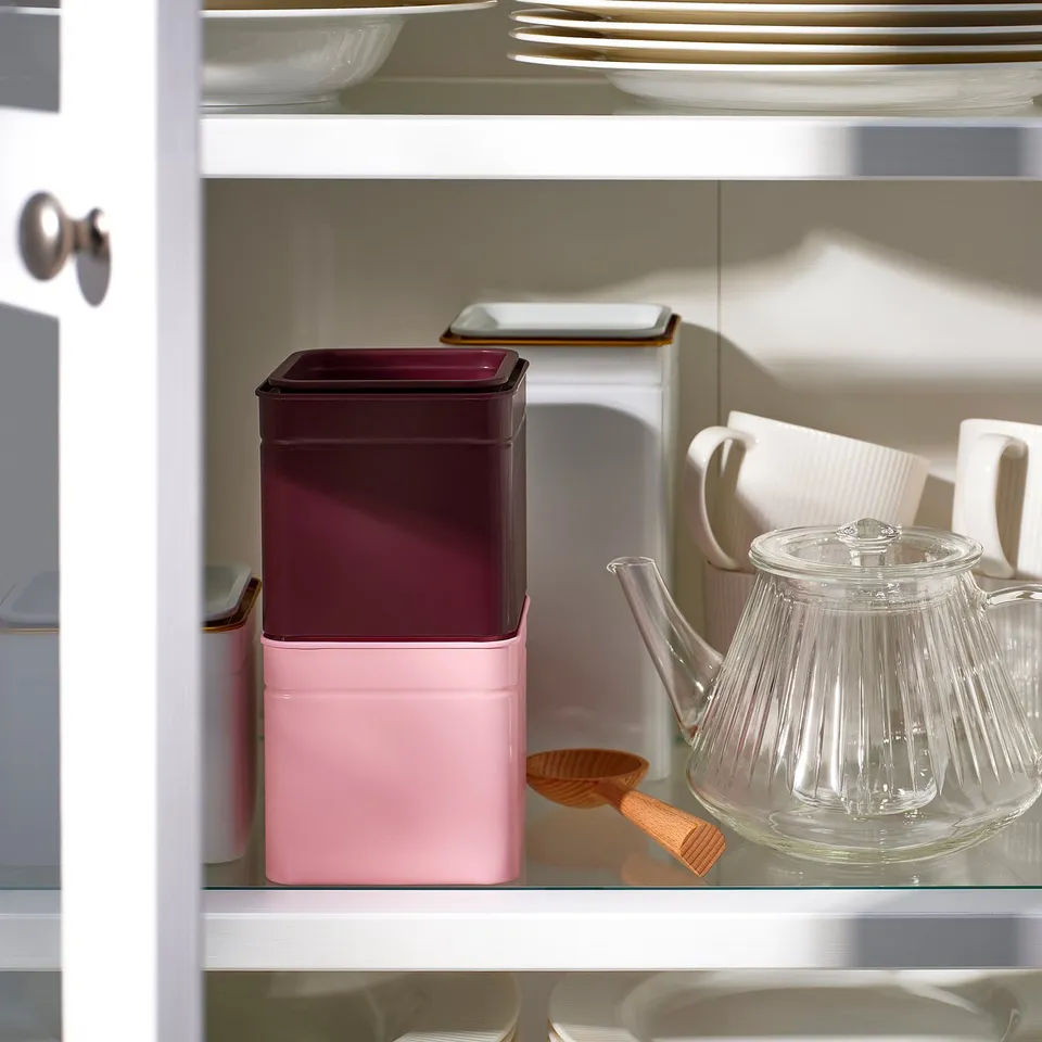 A kitchen shelf displaying a pink and maroon metal tin, a glass teapot, and ceramic mugs, ideal for storing coffee or tea.
