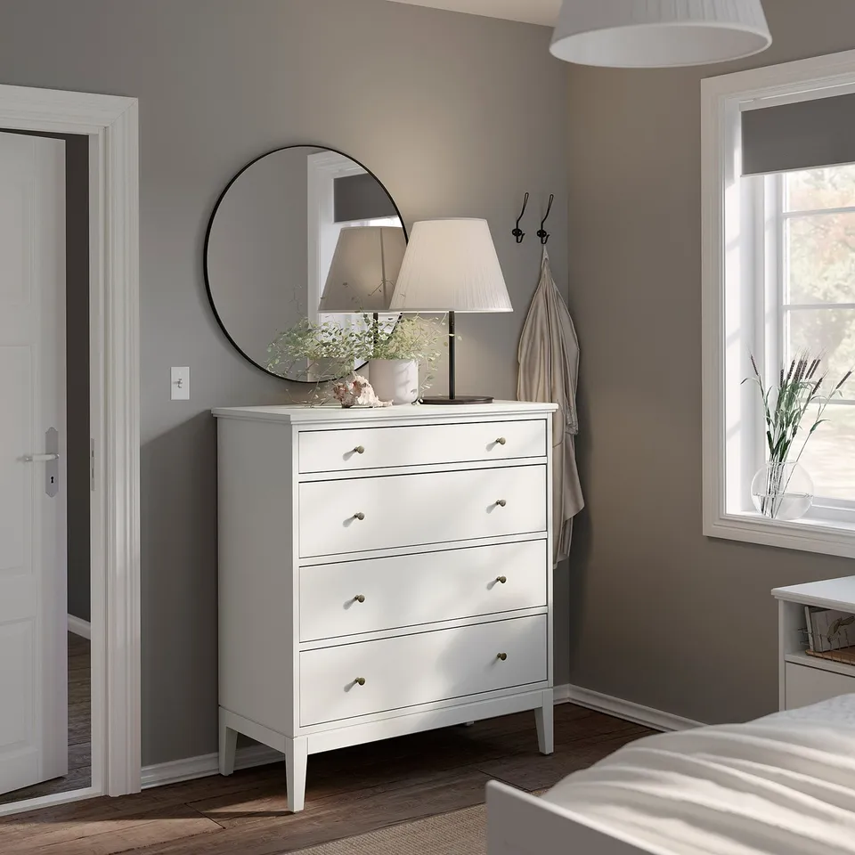 Modern white bedroom with round mirror, lamp, drawers, wooden floor, & window.