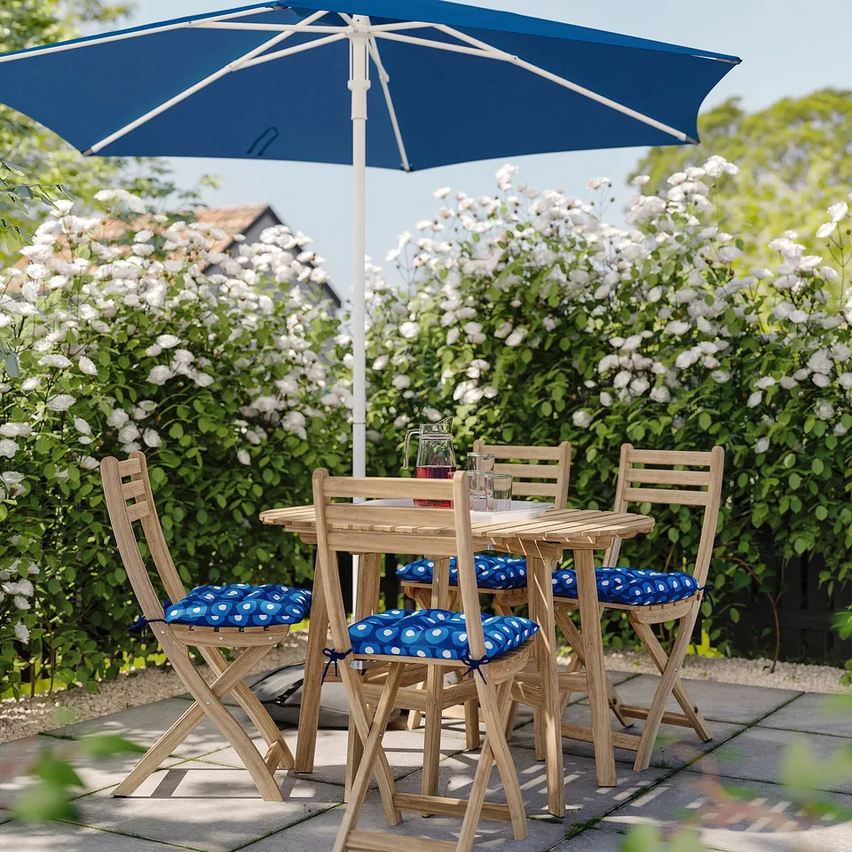 Garden patio with wooden furniture, cushioned chairs, blue umbrella, greenery, and white flowers.