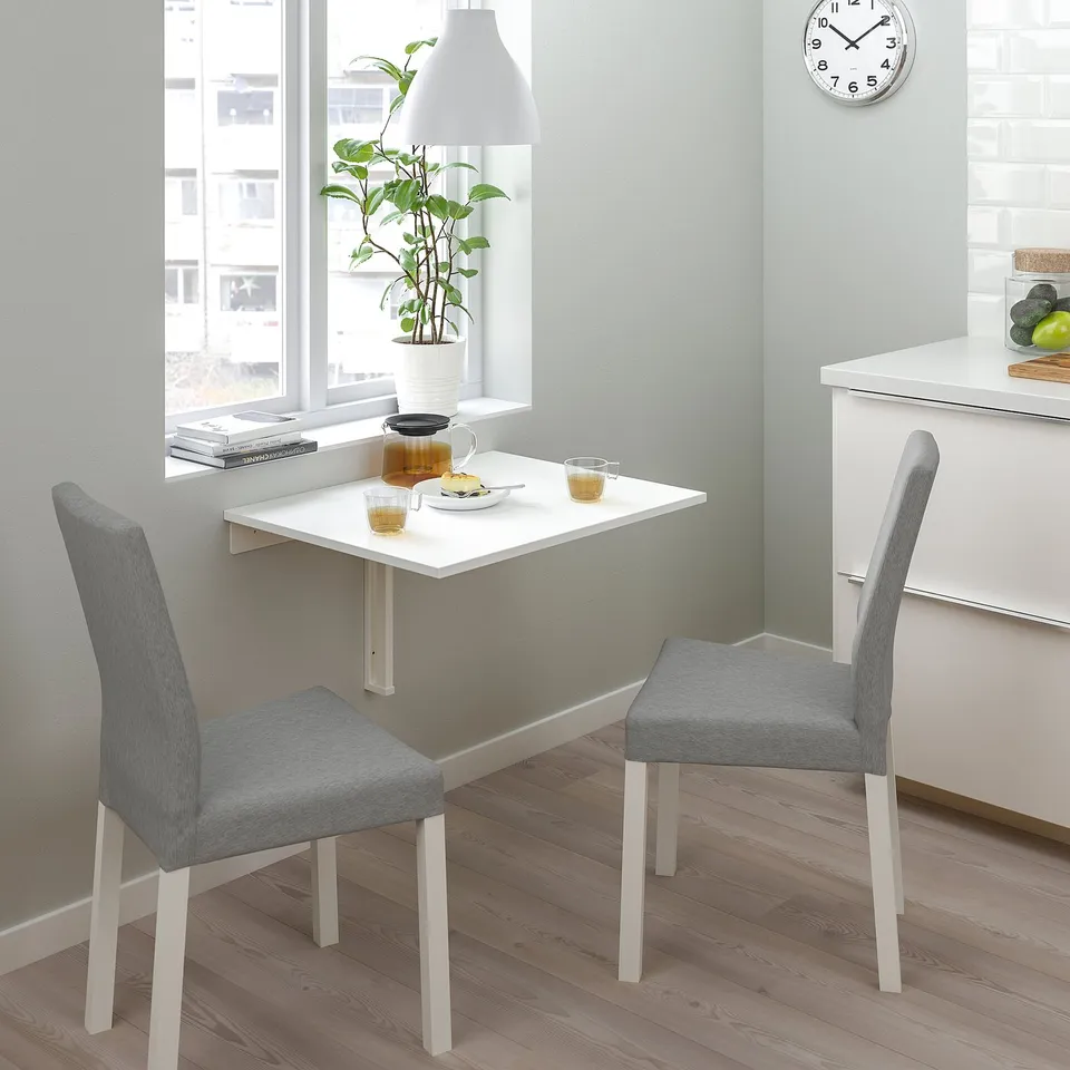 Small white wall-mounted drop-leaf table with two grey fabric NORBERG chairs, placed near a window with a clock above, creating a cosy breakfast nook.