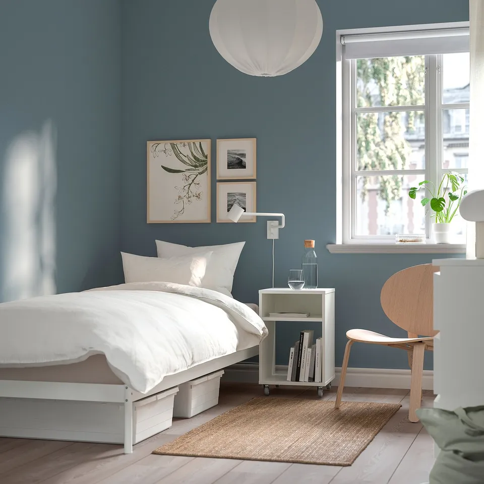 Modern blue bedroom with VEVELSTAD bed, chair, books, window, rug, and artwork. Clean and minimalist.