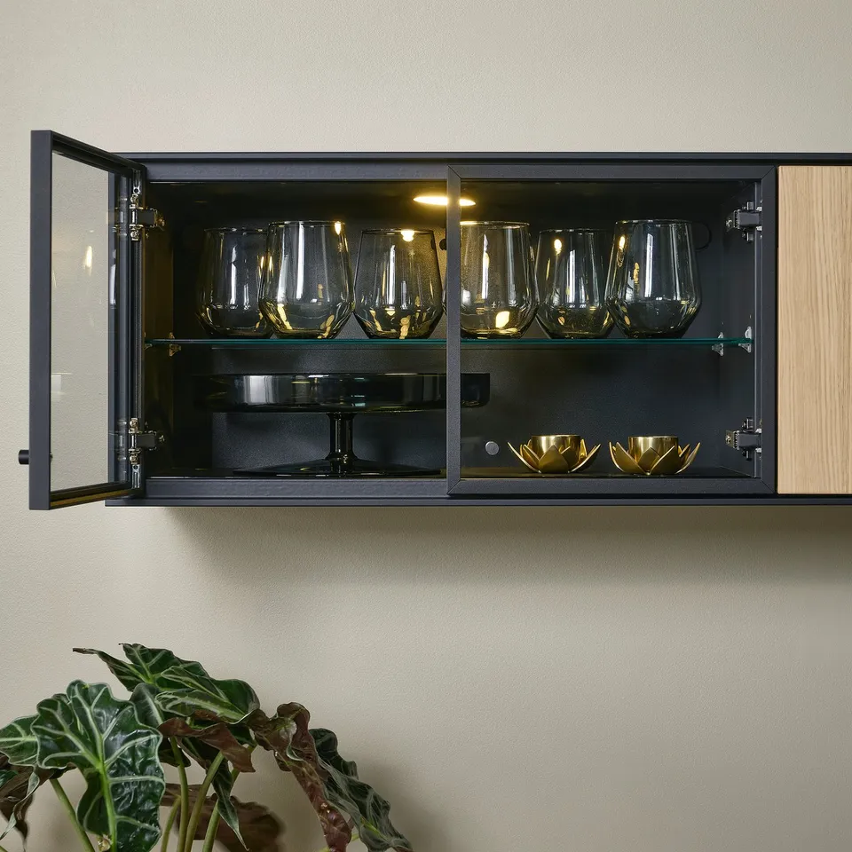 A wall-mounted display cabinet with glass doors, interior lighting, and four shelves showcasing glassware and decorative dishes.