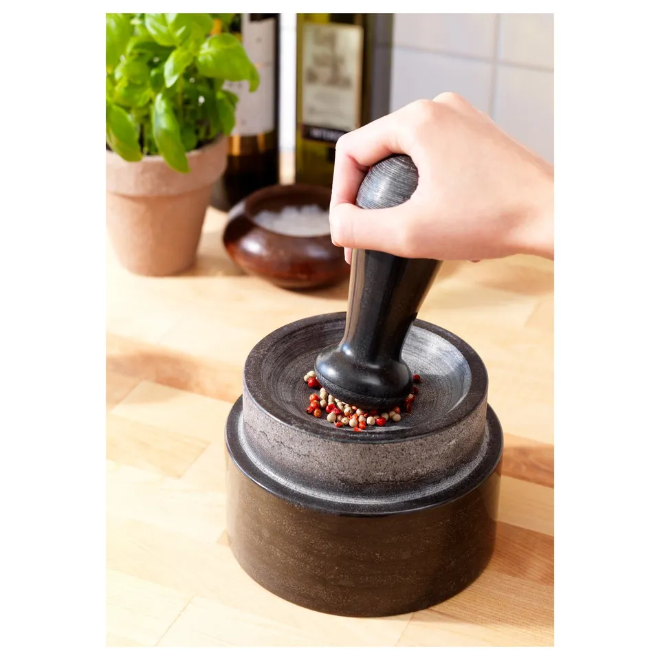 Hand presses pestle in marble mortar, crushing colourful spices on wooden table, with plant and bowl in background.