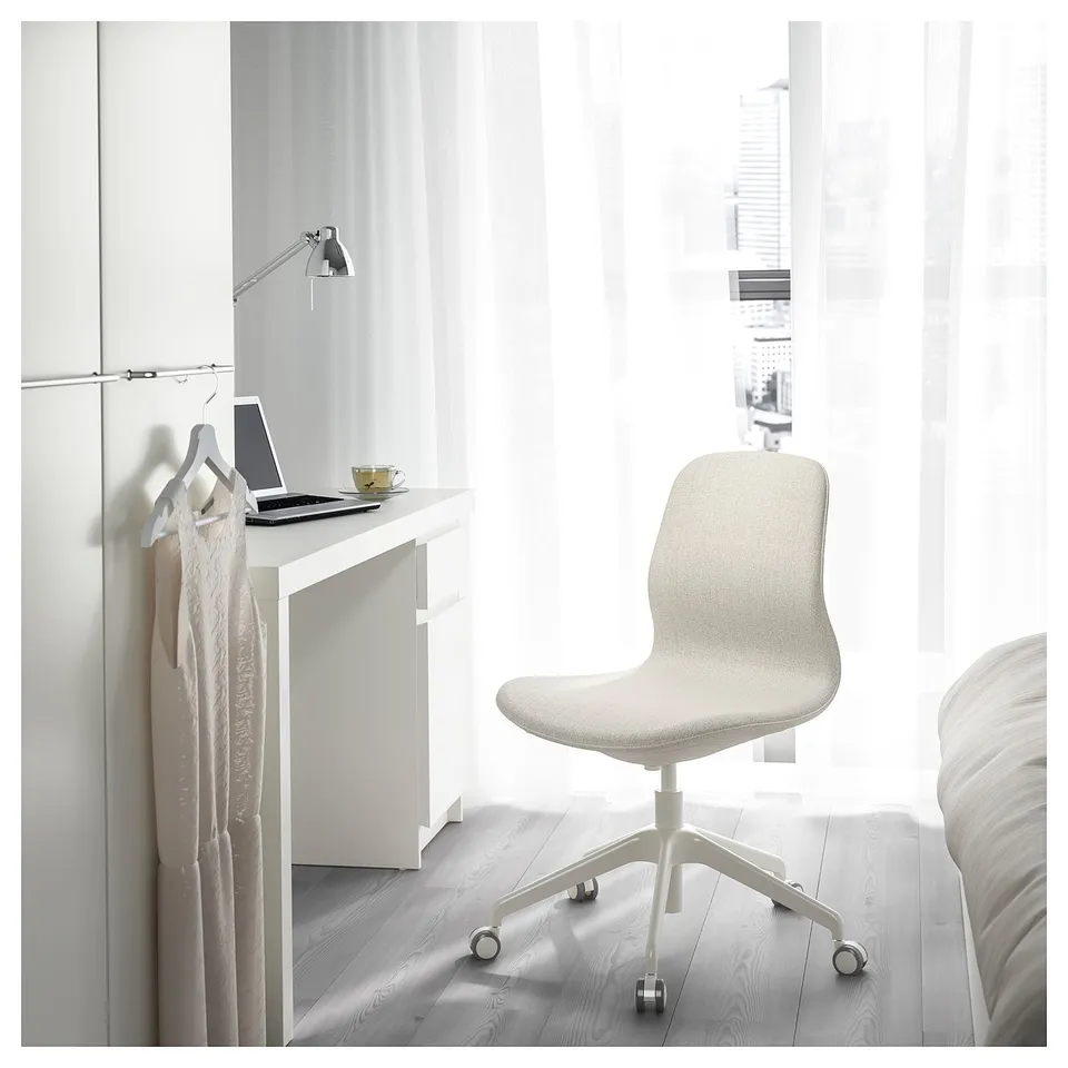 Minimalist home office setup with LÅNGFJÄLL beige fabric chair, white desk, laptop, cup, and clothes hanger, against window with white curtains.