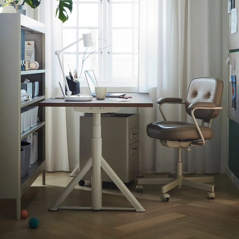 Modern home office setup with IDÅSEN adjustable desk, brown top, metal legs, and grey office chair by window.