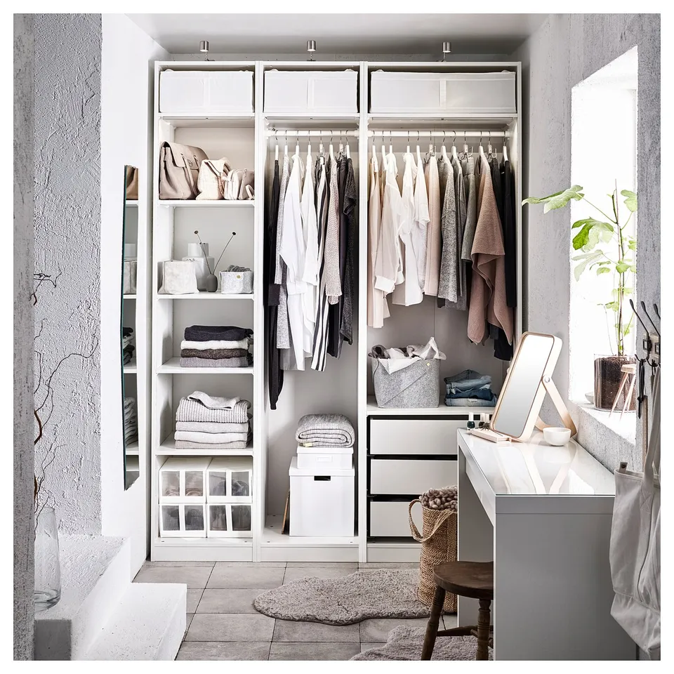 Organised closet with white PAX system. Hanging clothes, folded items, and storage bins. The room light-colored decor and a wooden chair.