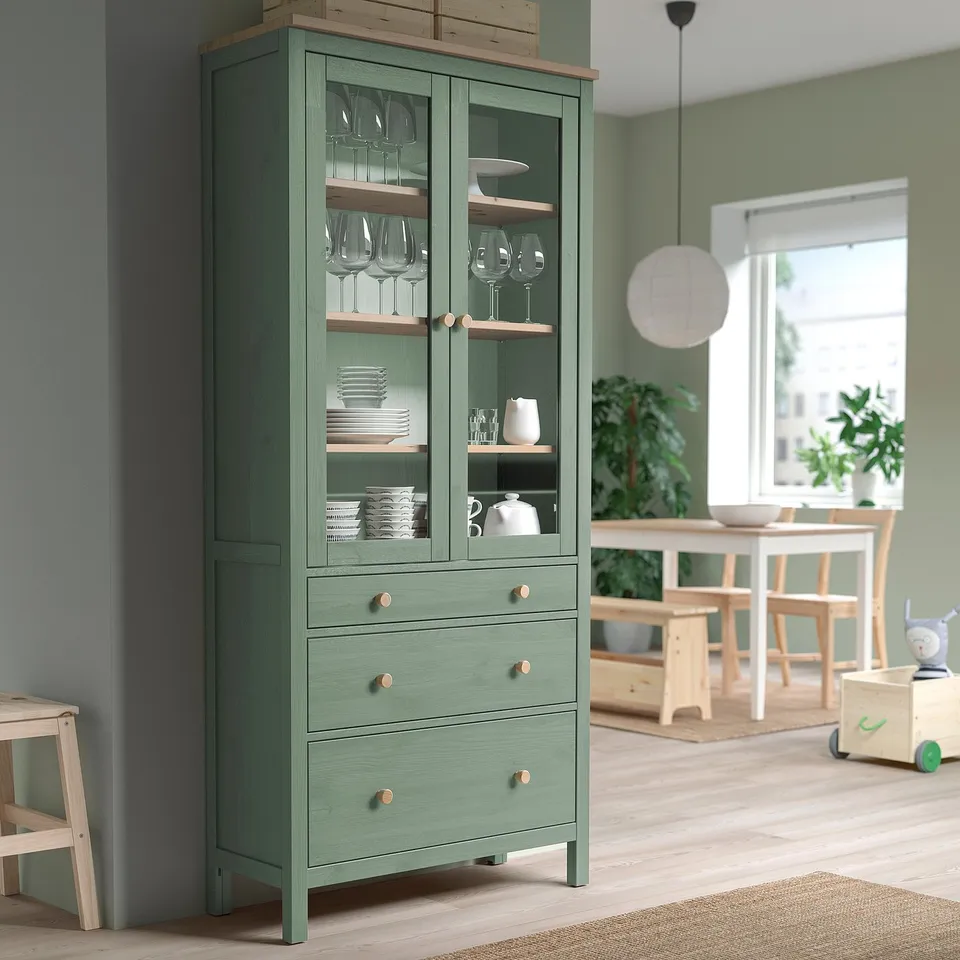 Green HEMNES cabinet with glass doors, holding wine glasses and plates, near dining area.