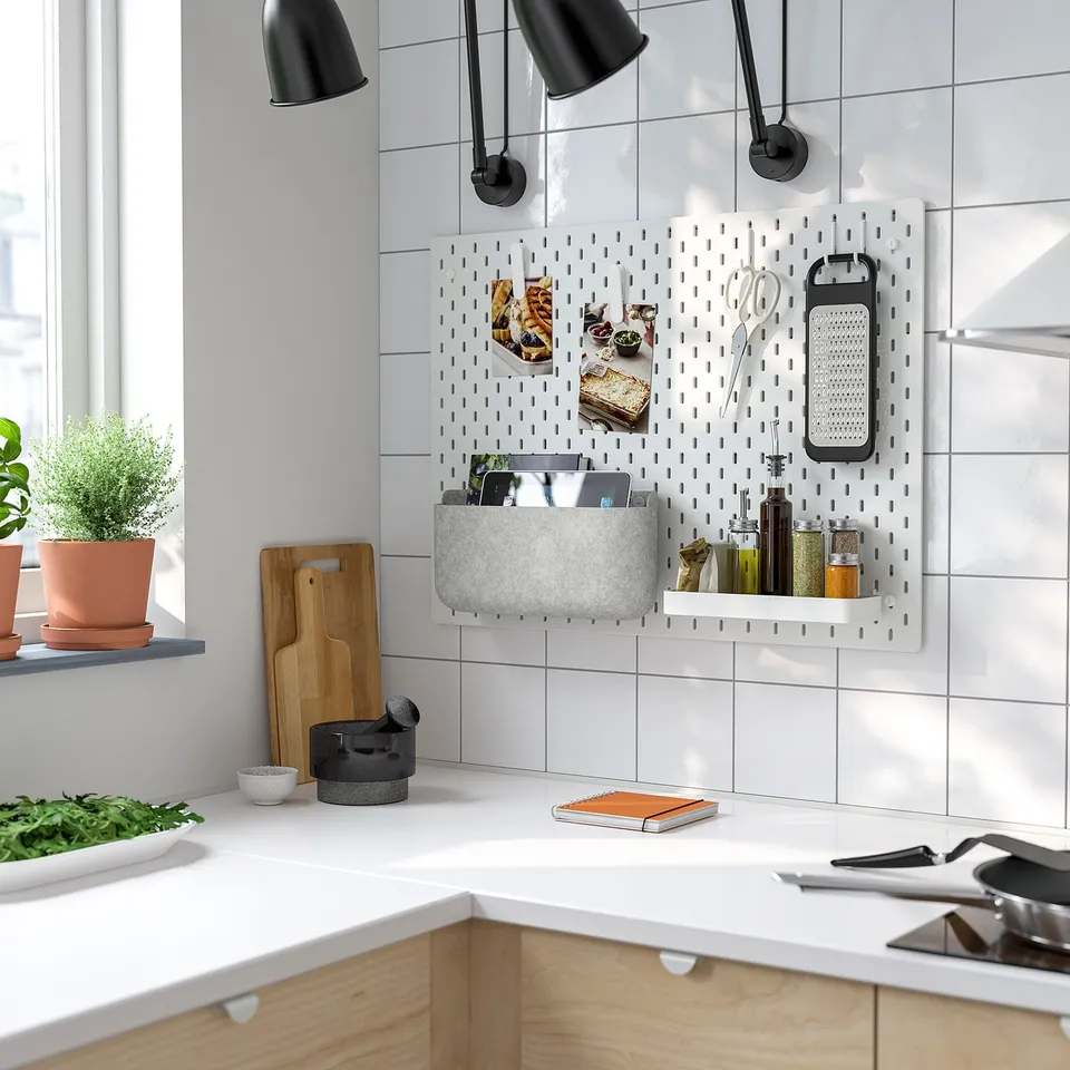 Modern white kitchen with SKÅDIS pegboard organiser holding utensils and spices near the window sill, accompanied by black lamps and wooden elements.