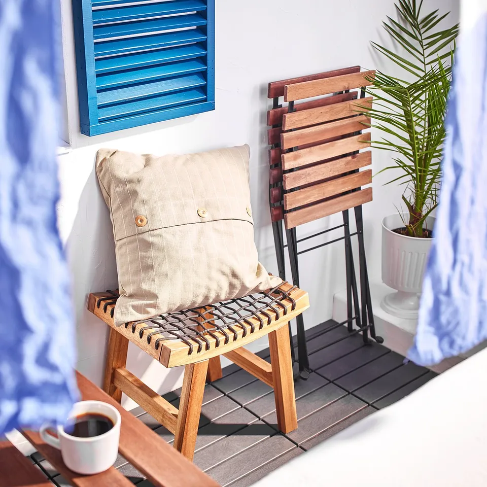 Balcony setup with STACKHOLMEN wooden stool, beige cushion, coffee on tray, potted plant near blue window shade.