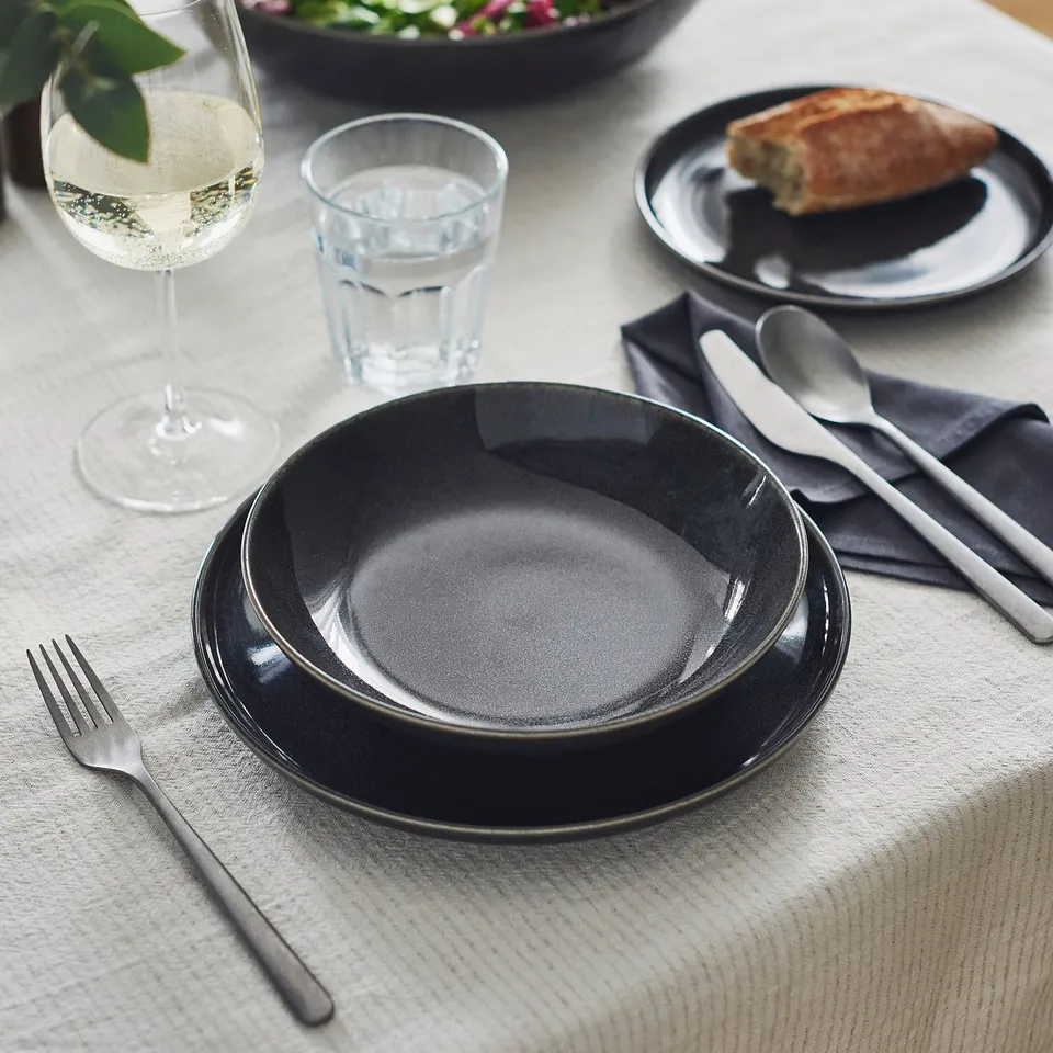 Elegant GLADELIG dark grey stoneware deep plate, medium-sized, on a neatly set table with cutlery, glass, and food. Handcrafted look.