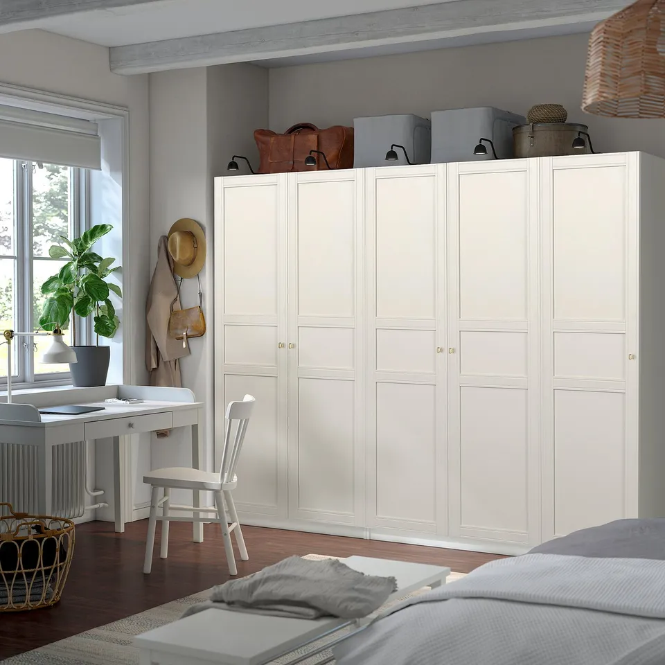 A spacious white PAX wardrobe, taller than a nearby desk and chair, storing various items atop its closed doors.