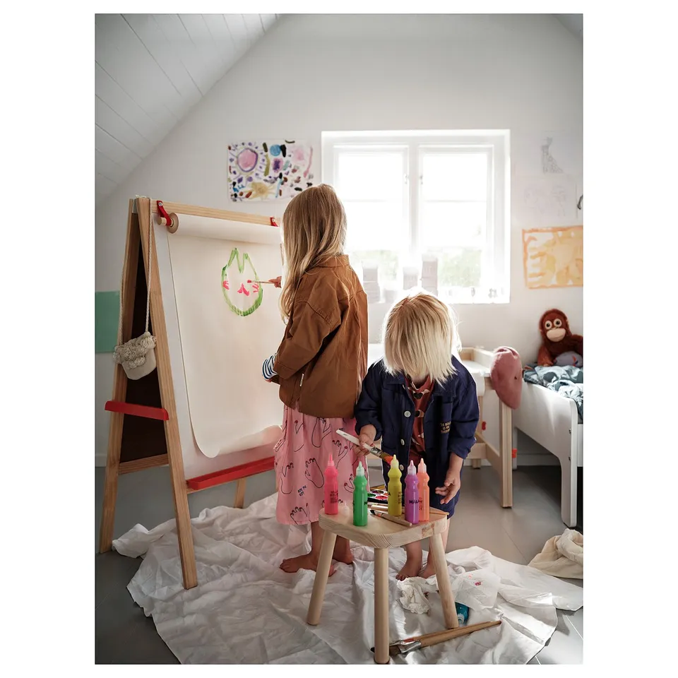 Two kids painting at MÅLA easel by window. One holds green bottle, other pink. Art on white paper, toys scattered, natural light.