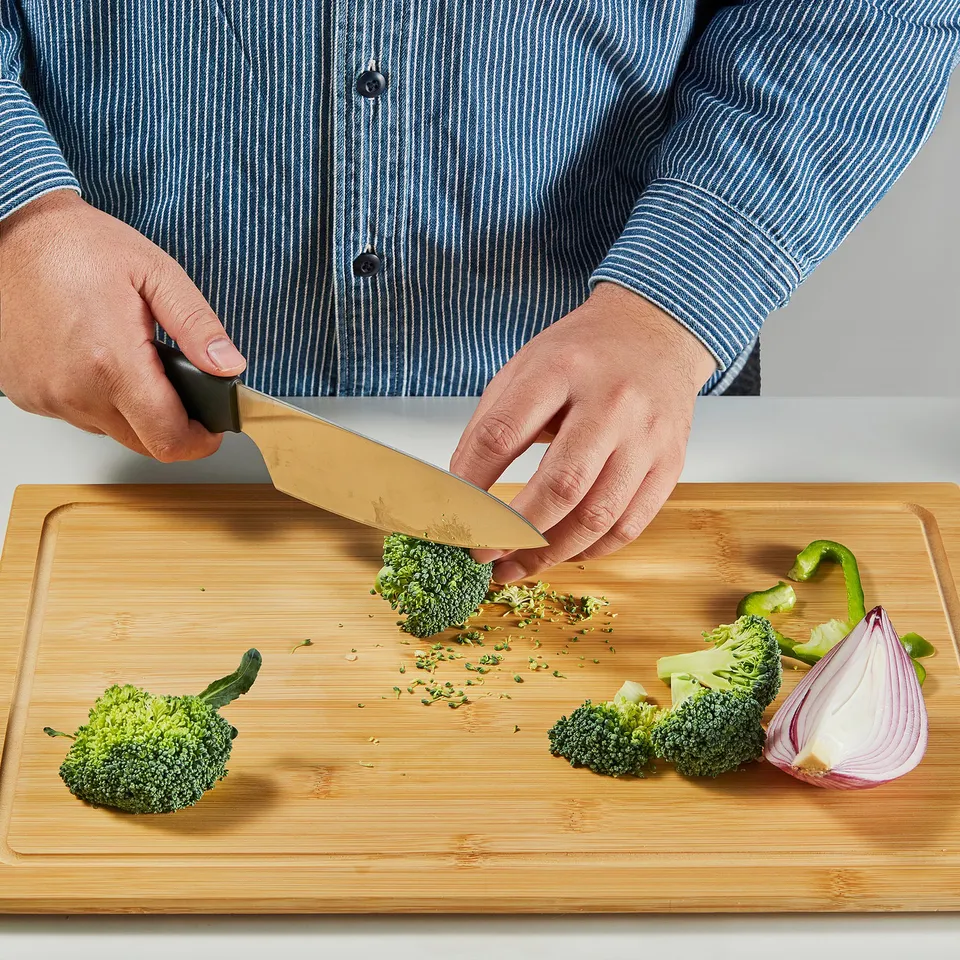 In a person is cutting broccoli on a bamboo cutting board using a cooks knife. The knife is durable and sharp, designed for everyday use.