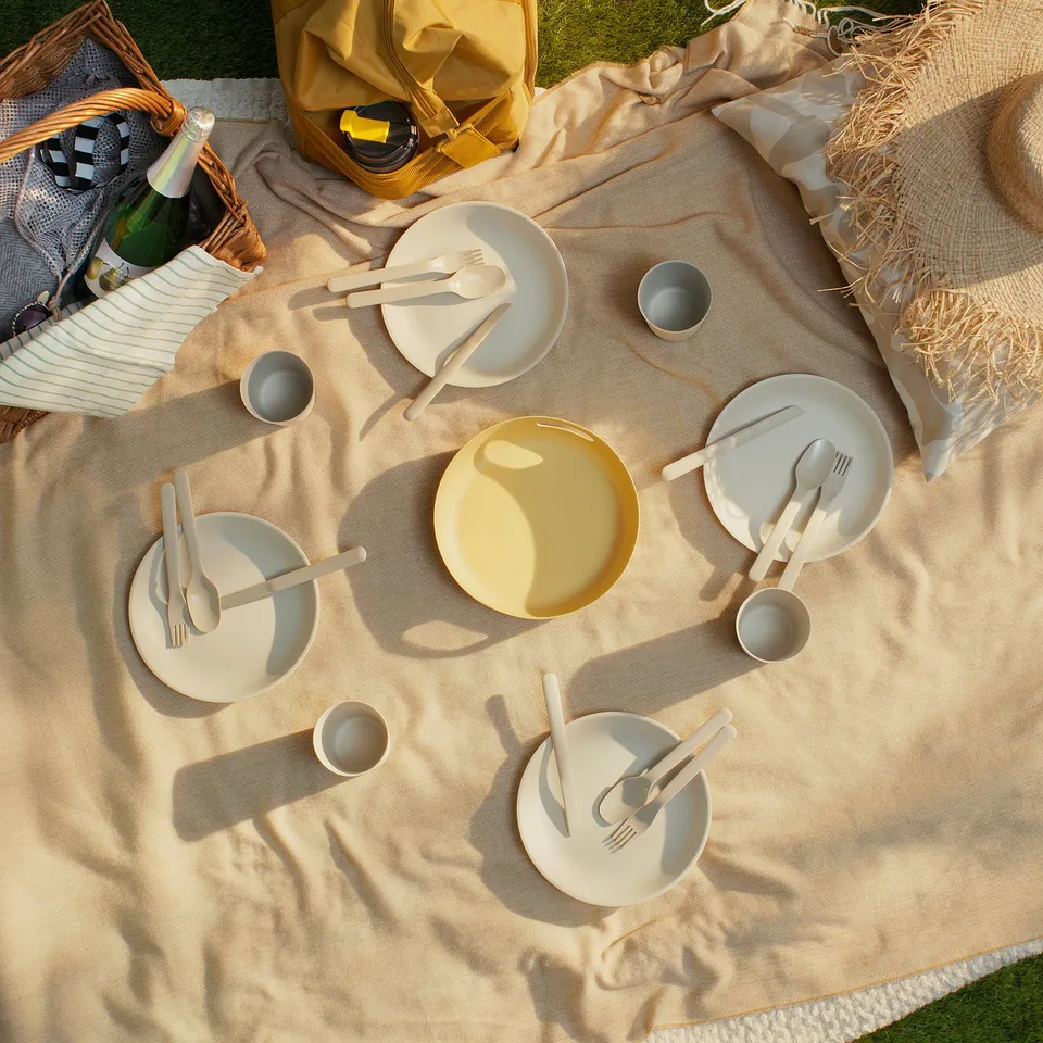 Picnic setup with beige plates, yellow bowl, cups, cutlery in beige bag on beige blanket.