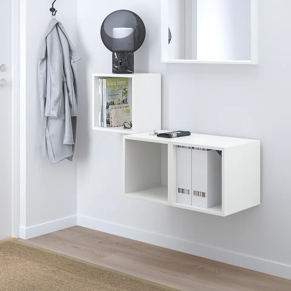 Minimalist white wall-mounted cabinets with open and closed storage, housing a round lamp, books, and a jacket on a hook.