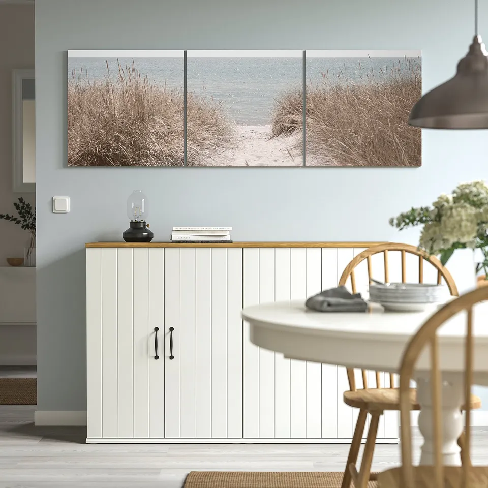 PJÄTTERYD beach photo trio on canvas, wooden frame, cabinet with lantern, books, and dining table with flowers.