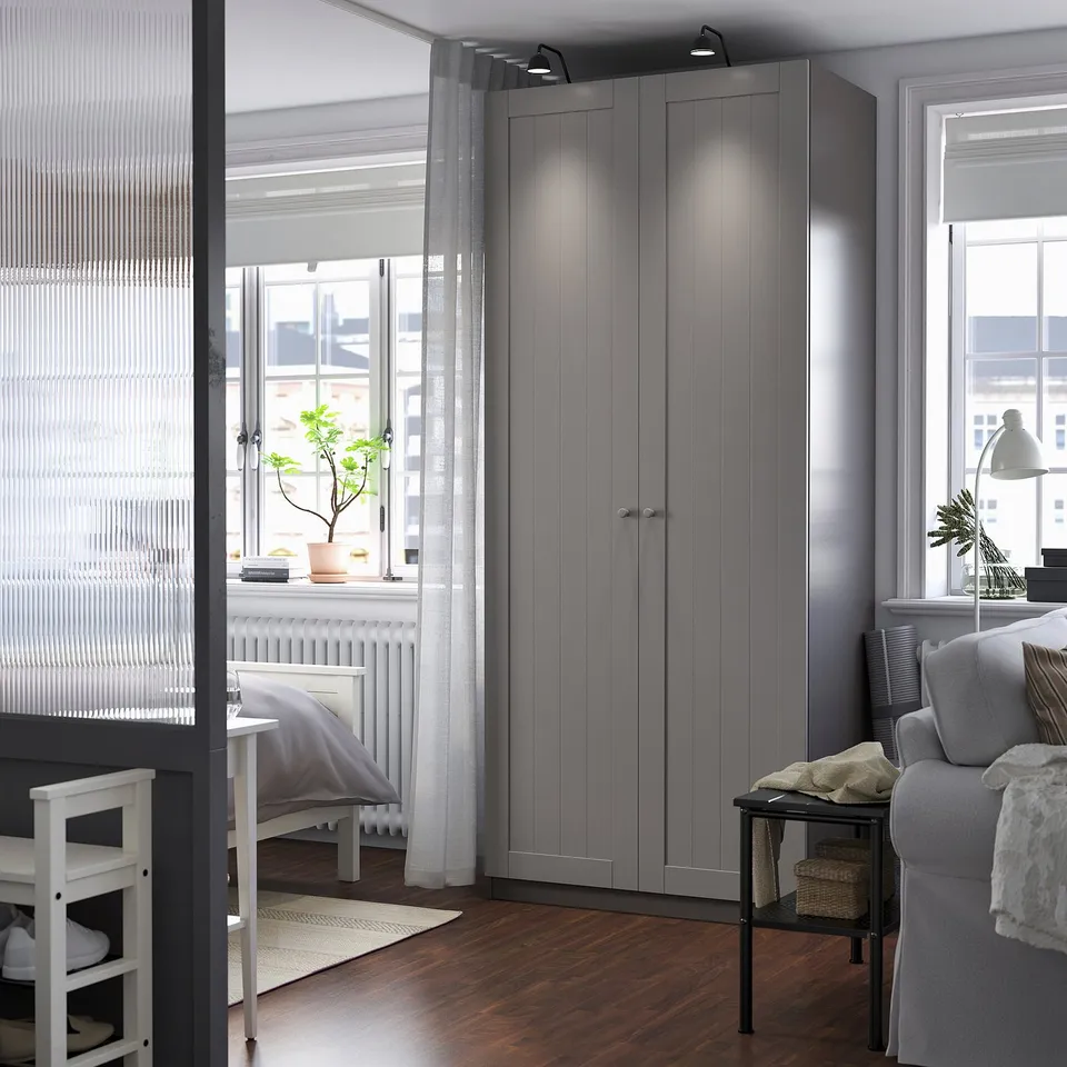 Modern grey PAX wardrobe, taller than desk, with three large doors, between window and sofa.