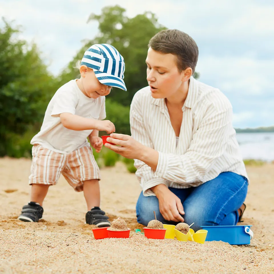Two people on a beach, one adult kneeling, using colourful sand moulds to play with sand, teaching a baby standing beside them.