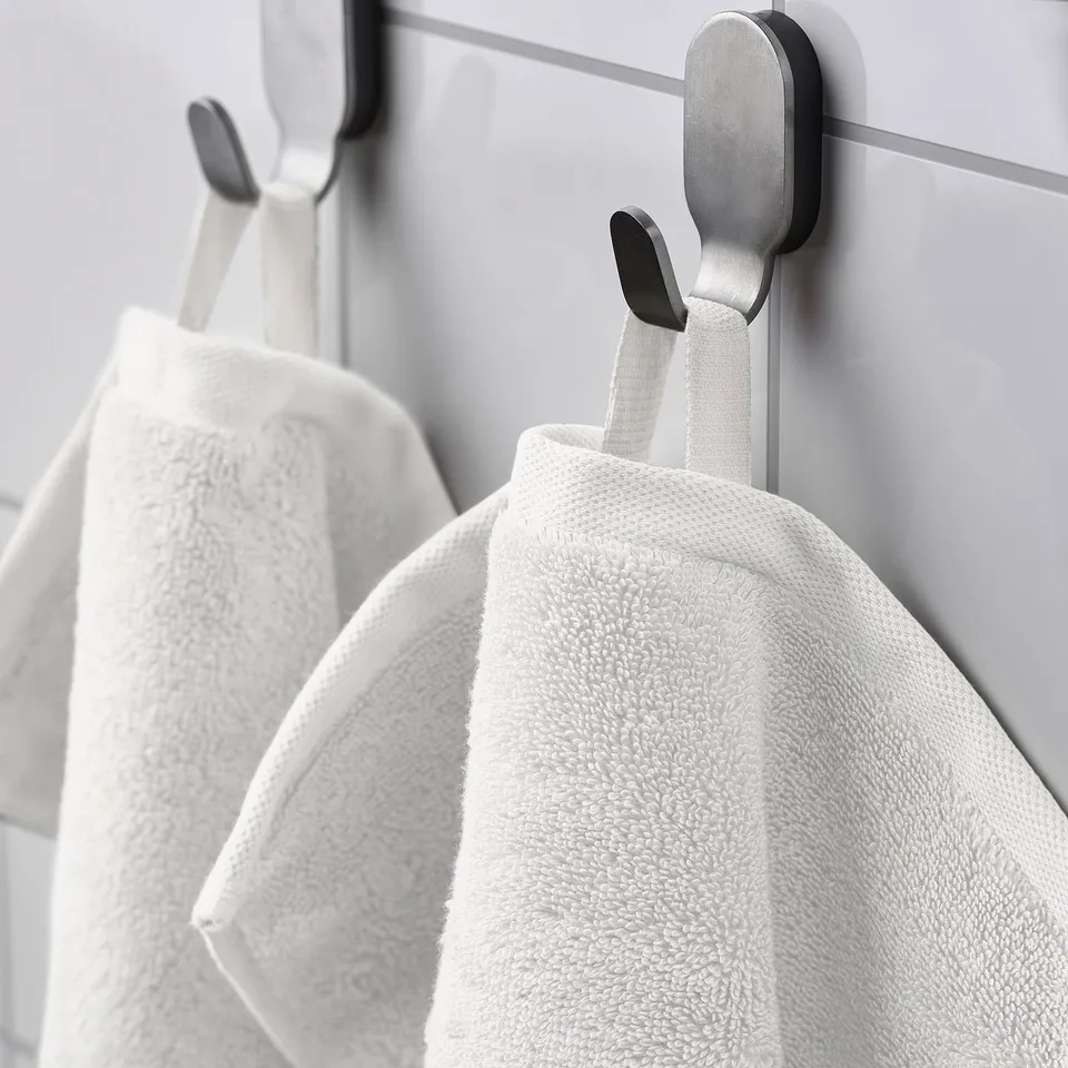 Three white towels hang neatly on silver and black hooks against a light grey wall. The hooks are part of a modern hanging system.
