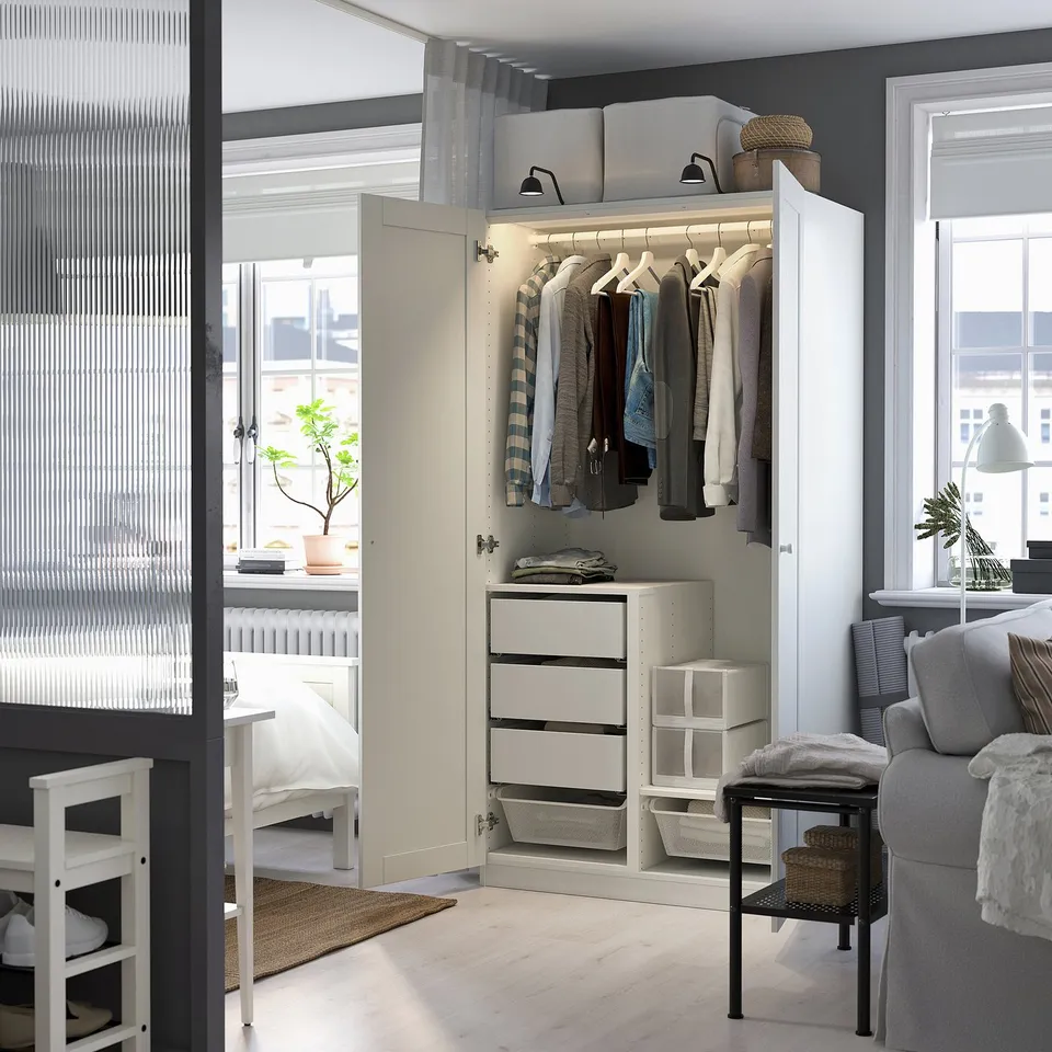 Modern white wardrobe with open space, lit inside, drawers, between bedroom and living room with chair, table, plant.