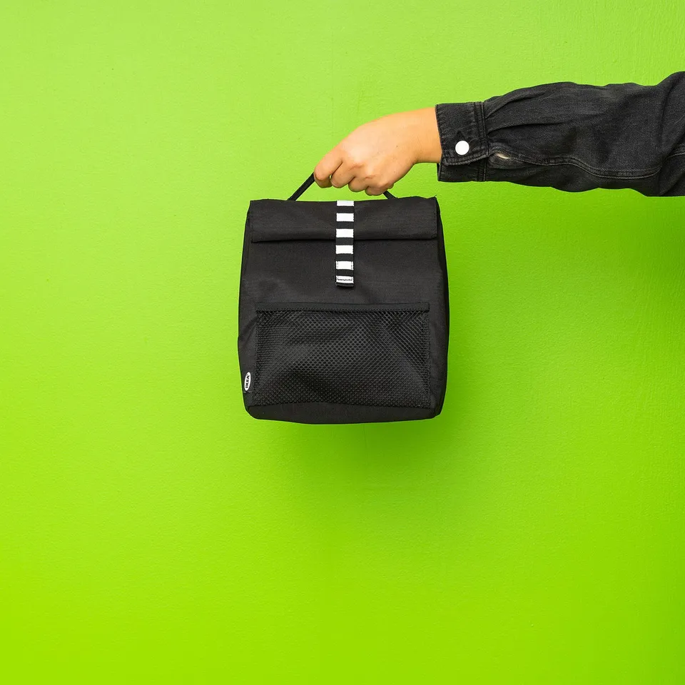 Person holding black rectangular lunch bag with mesh pocket against green wall.
