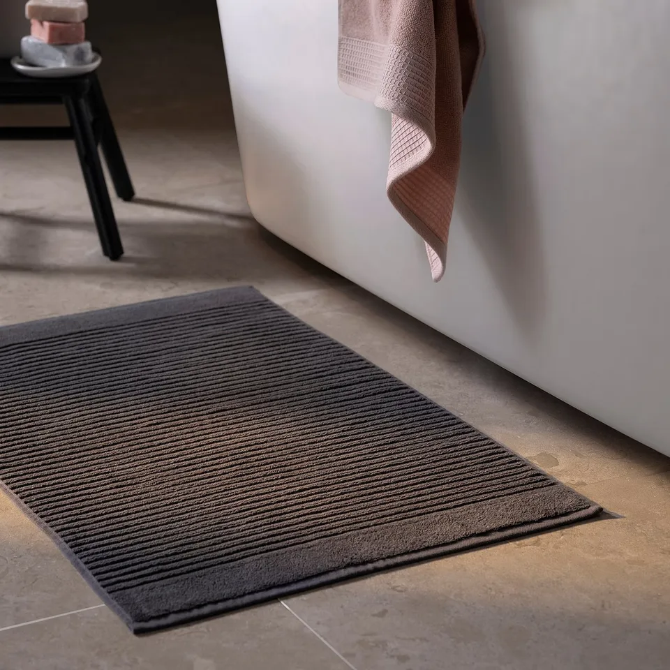 A dark grey bath mat named ALSTERN on a floor beside a tub. Its made of pure cotton, rectangular, and a ribbed texture.