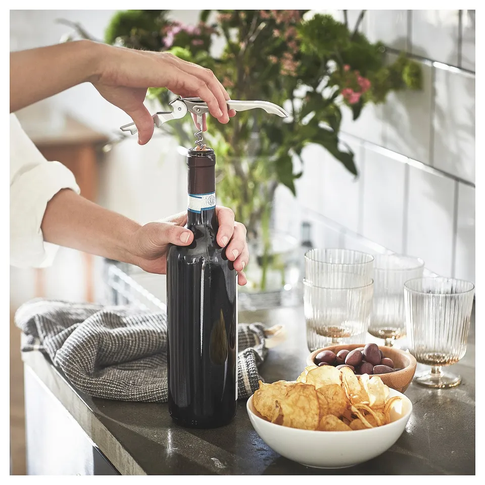 A person opens a wine bottle using a SEGELFISK stainless steel corkscrew on a counter with snacks and glasses.