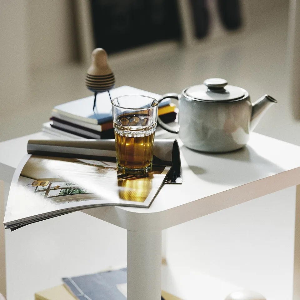 A white table holds a glass teacup with warm tea, a silver teapot, and an open magazine.