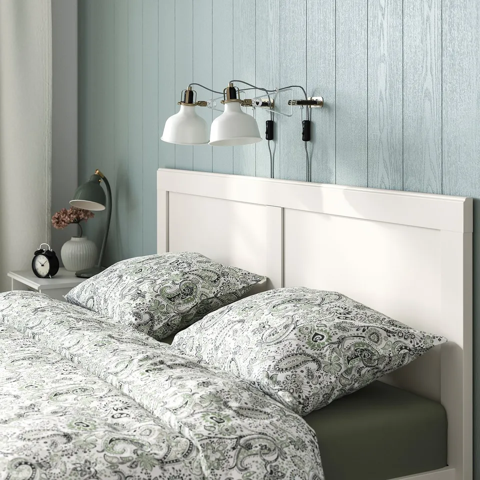 Bed with floral sheets, white headboard, two lamps, vase with flowers, and an alarm clock on bedside table.