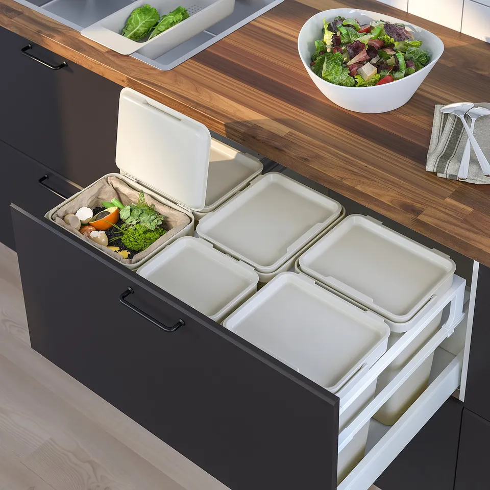 Modern kitchen with waste sorting bins. Dark cabinets, white recycling bins. One open bin with veg scraps. Wooden counter, salad bowl.