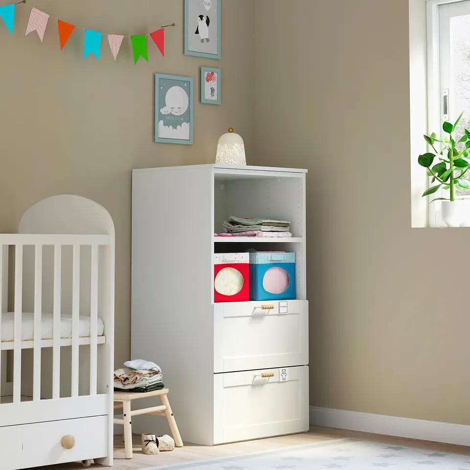 Small white nursery with baby crib, low stool, and storage unit. Colourful decor and books.