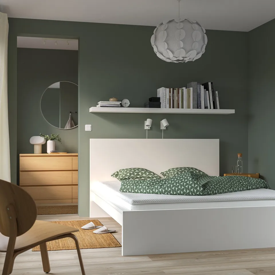 Modern bedroom: large white bed with green pillows, wooden chair, round mirror. Green walls, floating shelf with books.