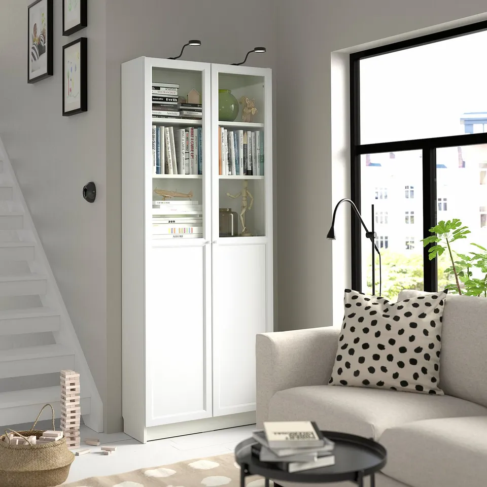 White BILLY bookcase with glass doors in living room, holding books, decor, lamp. Nearby beige sofa with polka dot pillow.