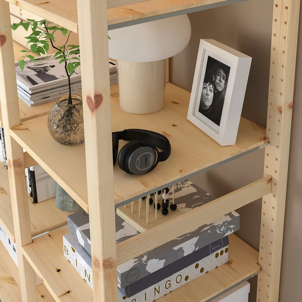 Wooden shelf unit with adjustable shelves, holding various items including headphones, picture frame, lamp, books, and plant.