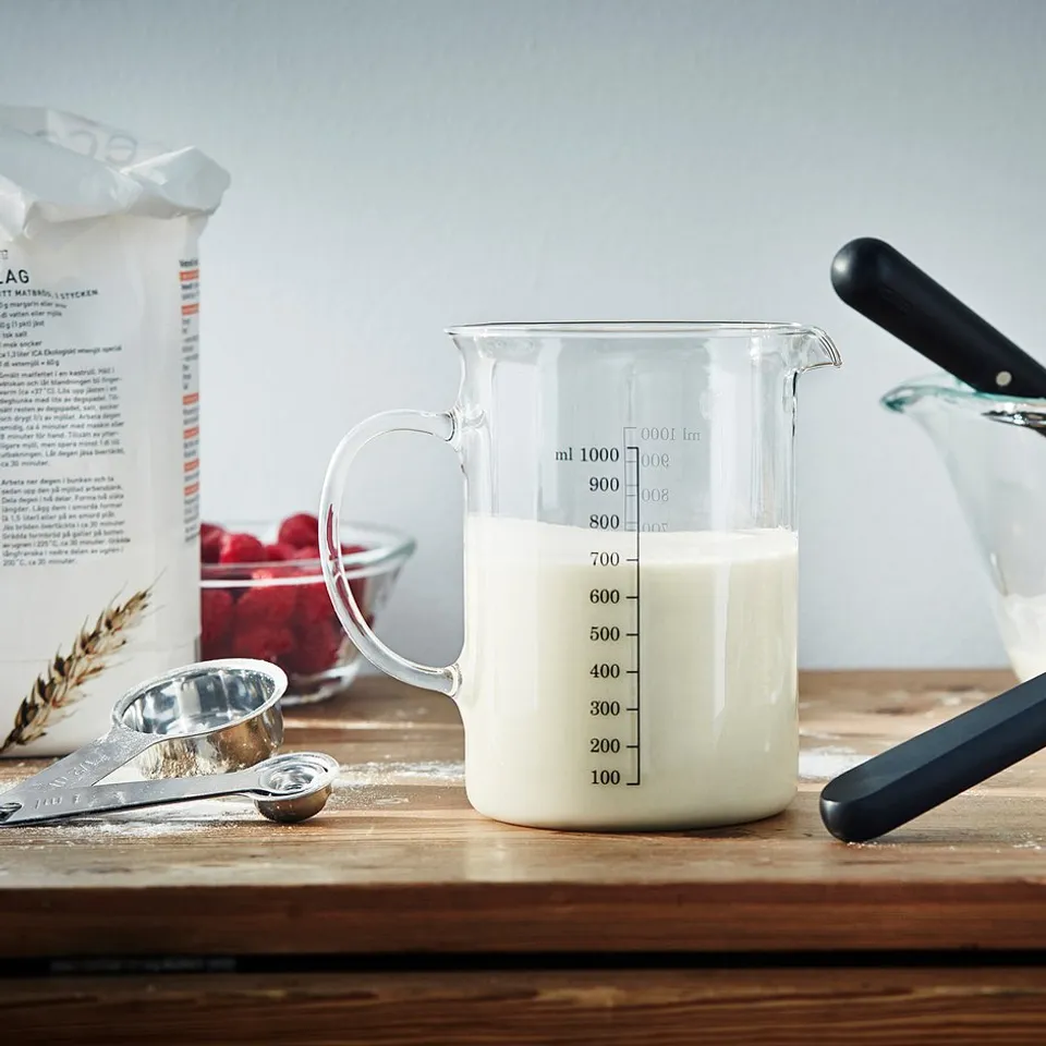 Clear glass measuring jug on wooden table with flour bag, measuring spoons, crepes, and phone.
