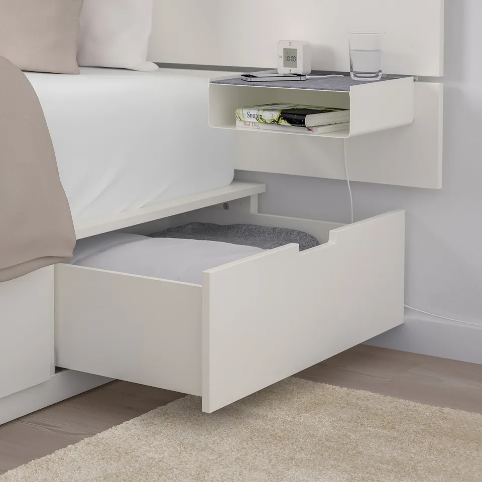 Bedside unit with open shelf displaying books and a glass, under-bed drawer open showing storage.