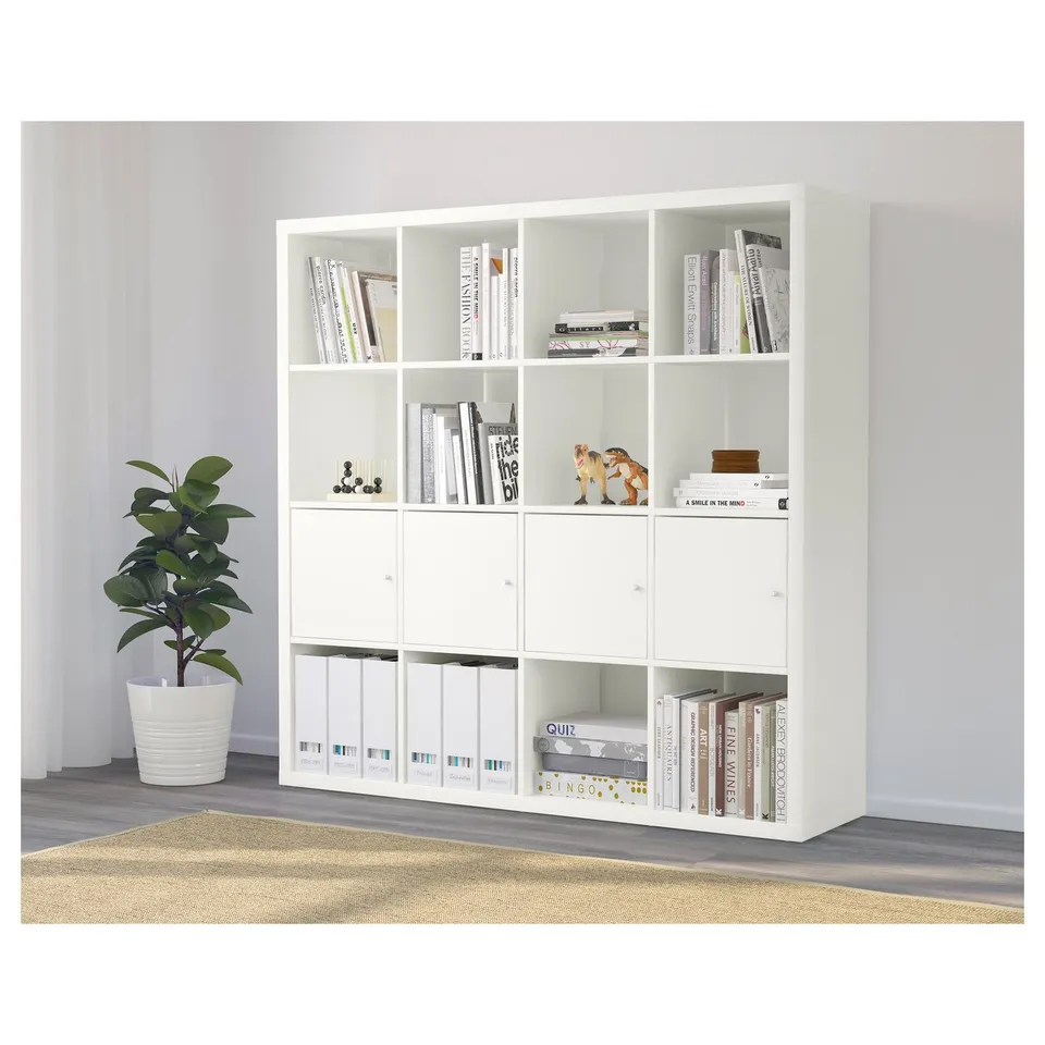 White KALLAX shelving unit holding books, decorative items, and filing folders, with a potted plant nearby.