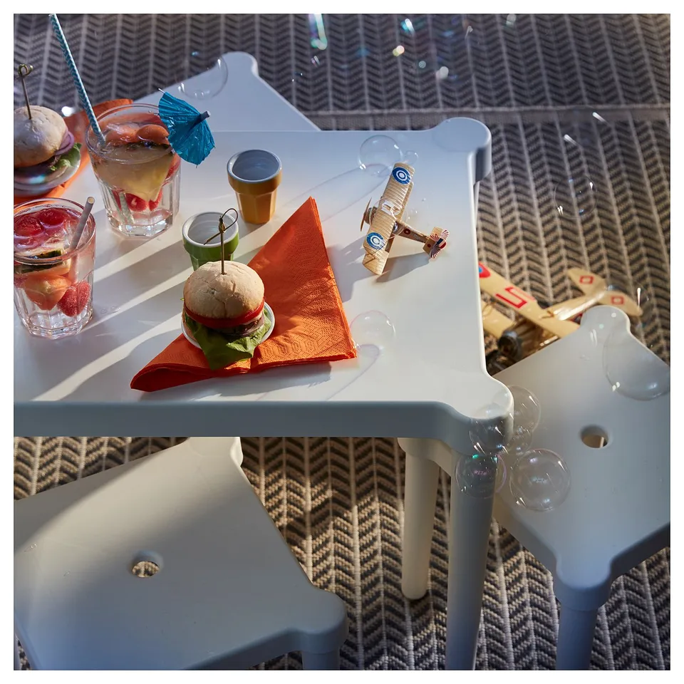Childrens play table with stackable white stools. Mini sandwiches and drinks on table. Wooden toy plane and farm vehicle nearby.