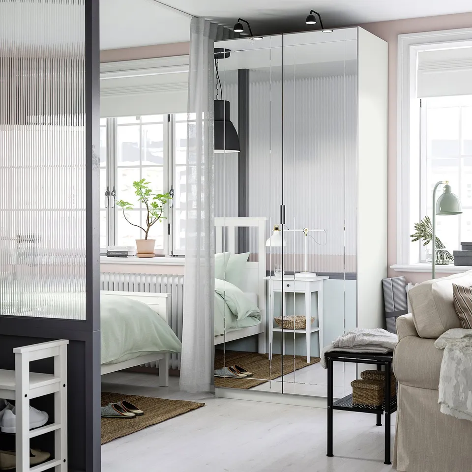 Modern minimalist room with large PAX wardrobe, mirrored sections, cosy bed, lamp, couch, and a plant on the window sill.