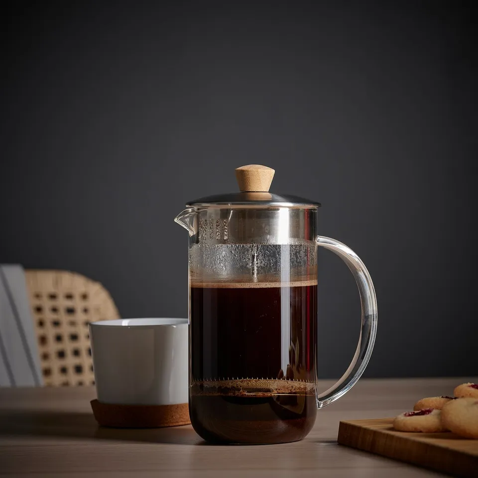 Clear glass coffee/tea maker with wooden handle and strainer, brewing dark liquid, accompanied by a white mug and cookies on a wooden surface.