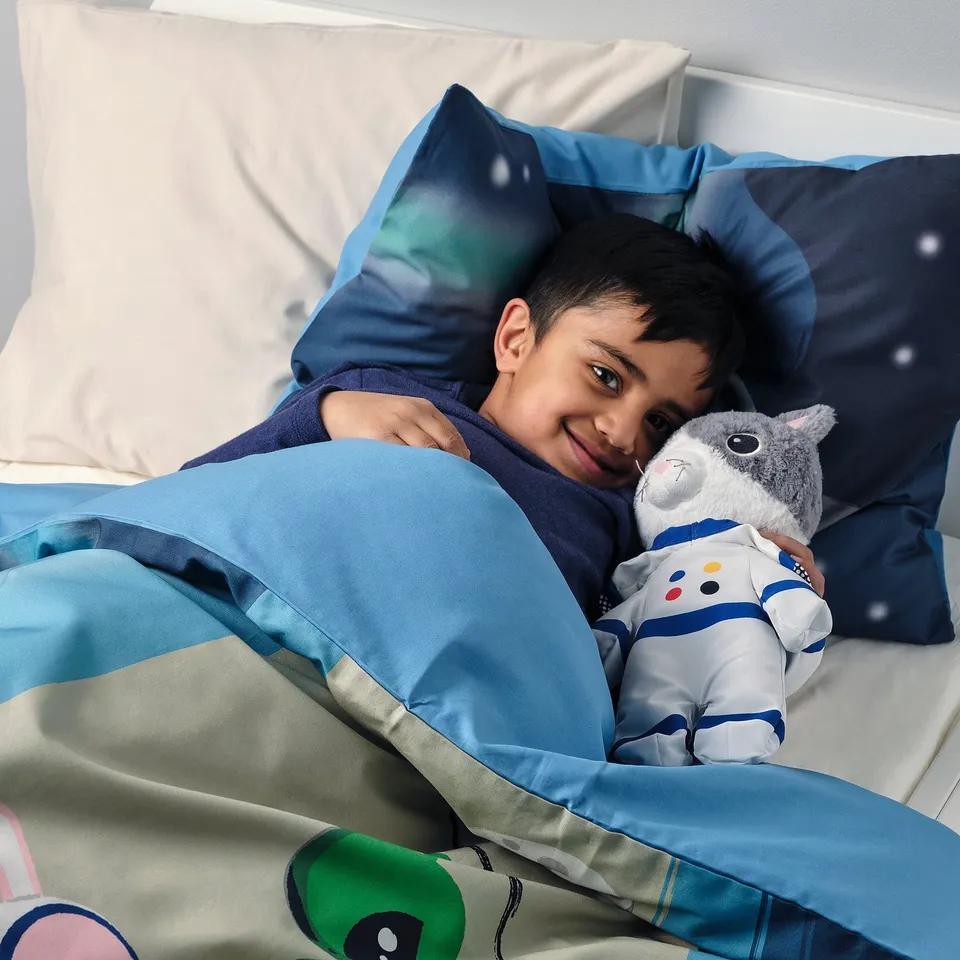 Happy child lying on bed, hugging blue and white astronaut cat cuddly toy, using star-patterned pillow.