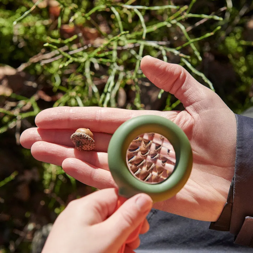 Hands hold small compass and magnifying glass outdoors. Compass has green ring, and magnifying glass shows detailed texture.