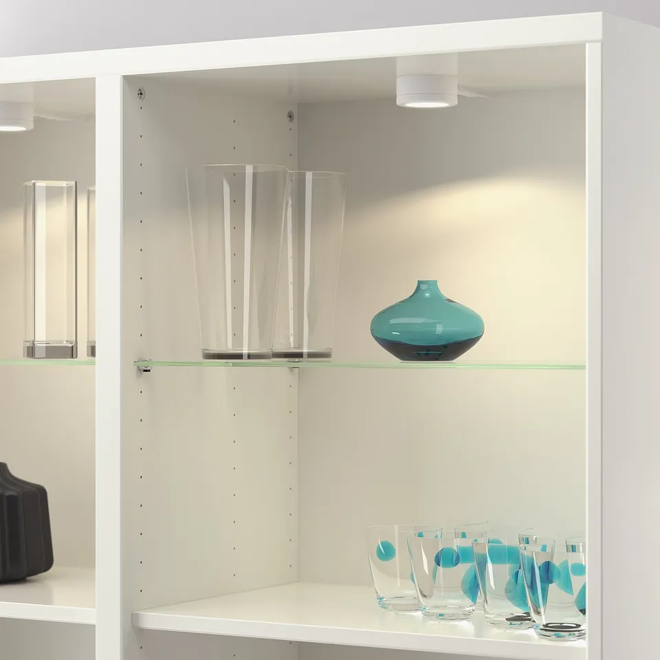 White shelf unit with glass shelves, displaying clear and blue glass vases and cups.