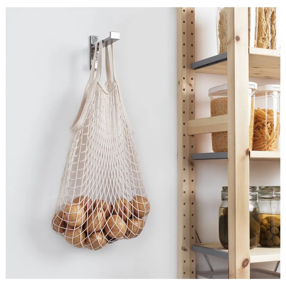 Mesh produce bags hang on wall hook in kitchen, storing potatoes beside wooden shelving units with food items.