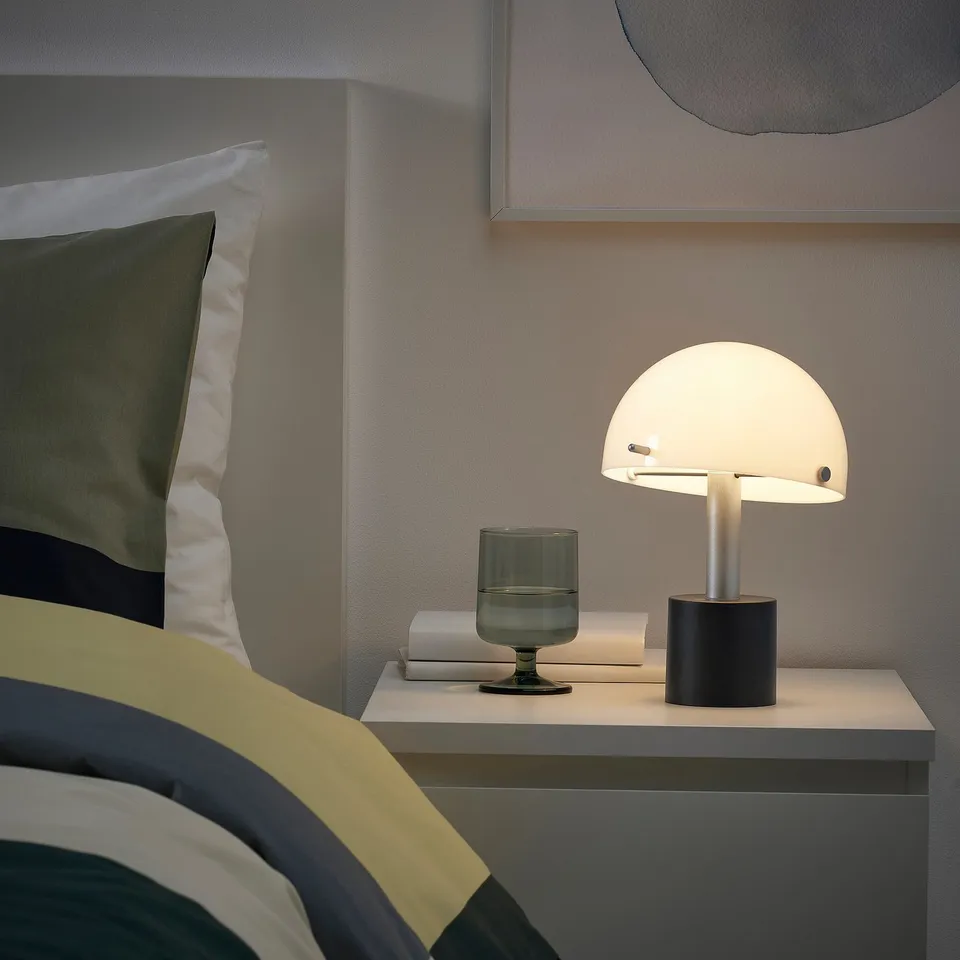 Small, stylish lamp on bedside table with soft light, grey base, white shade, green vase, book. Cosy room with striped bedding.