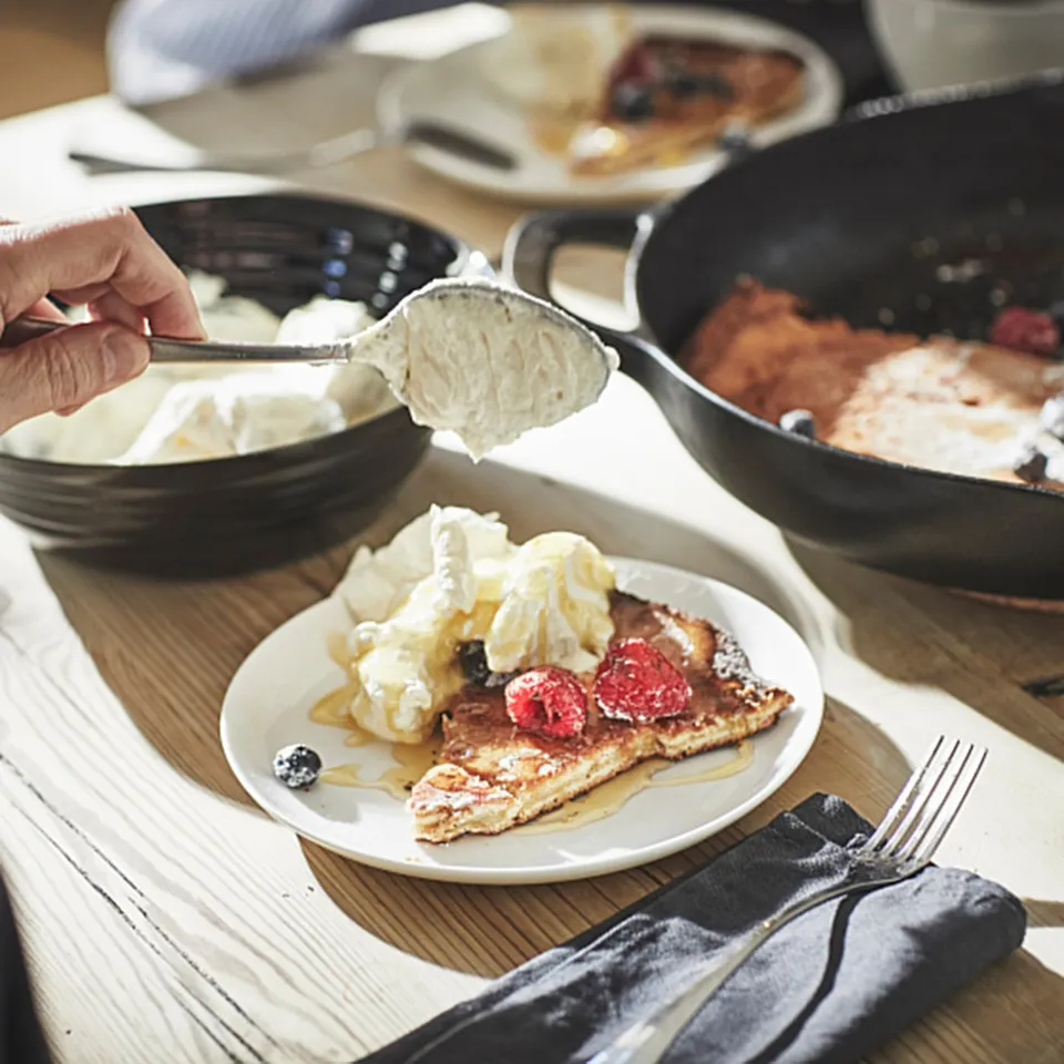 A person serves whipped cream onto french toast with berries and syrup on a white plate, with cutlery and other dishes in the background.