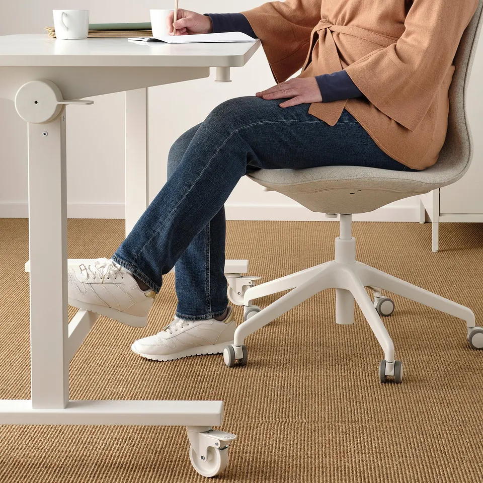 Person sitting at adjustable table with footrest, wearing jeans, trainers, and an apron. They are writing with a pen and a cup is on the table.