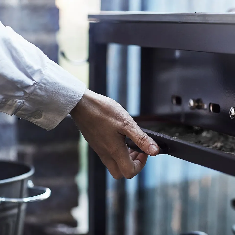 A person adjusts a GRILLSKÄR grills lid, which is silver and made of stainless steel. The grill has built-in controls and a thermometer hood.