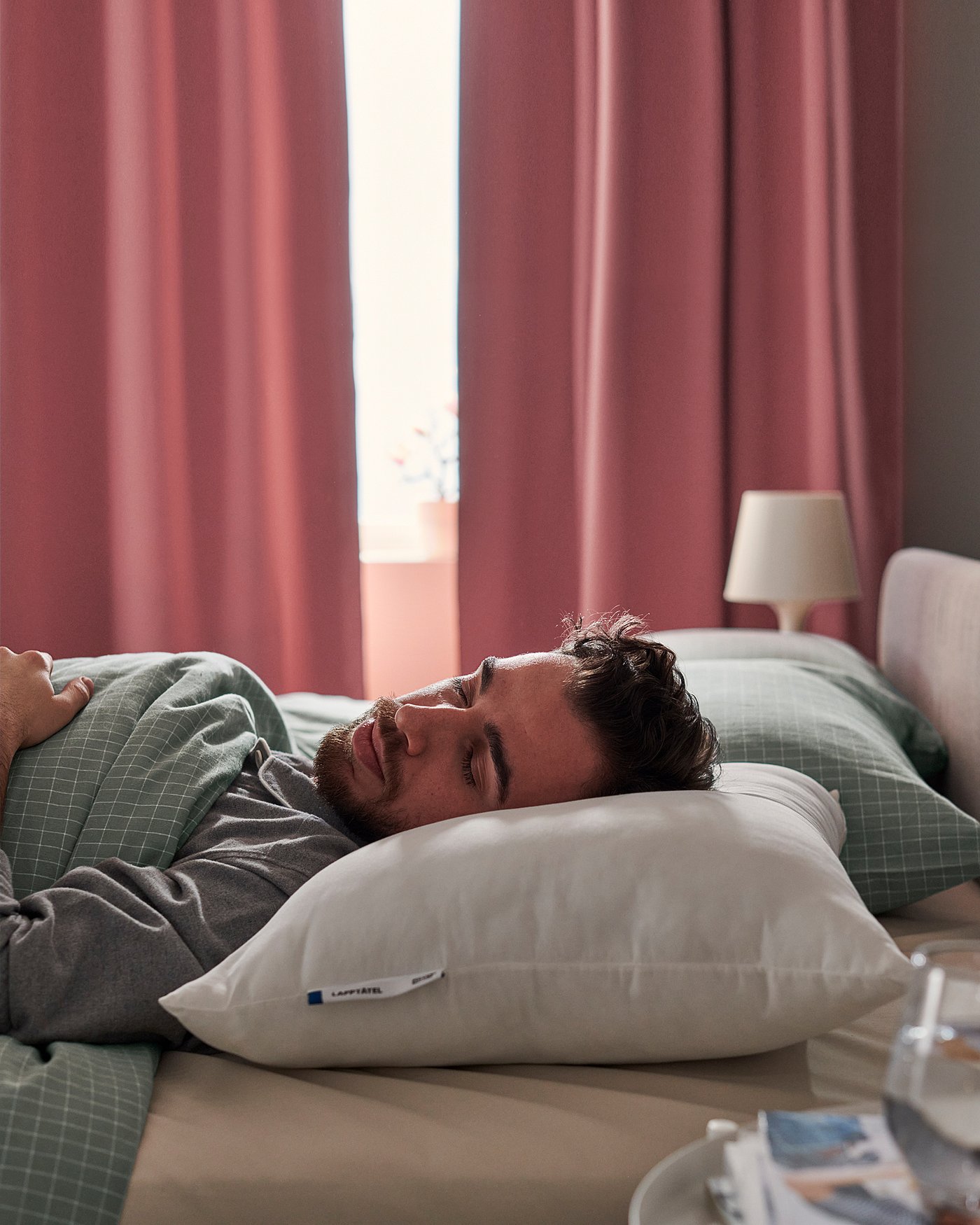 Person sleeping on a white LAPPTÅTEL pillow in a bed with green and white linens, red curtains, and a bedside table with a lamp.