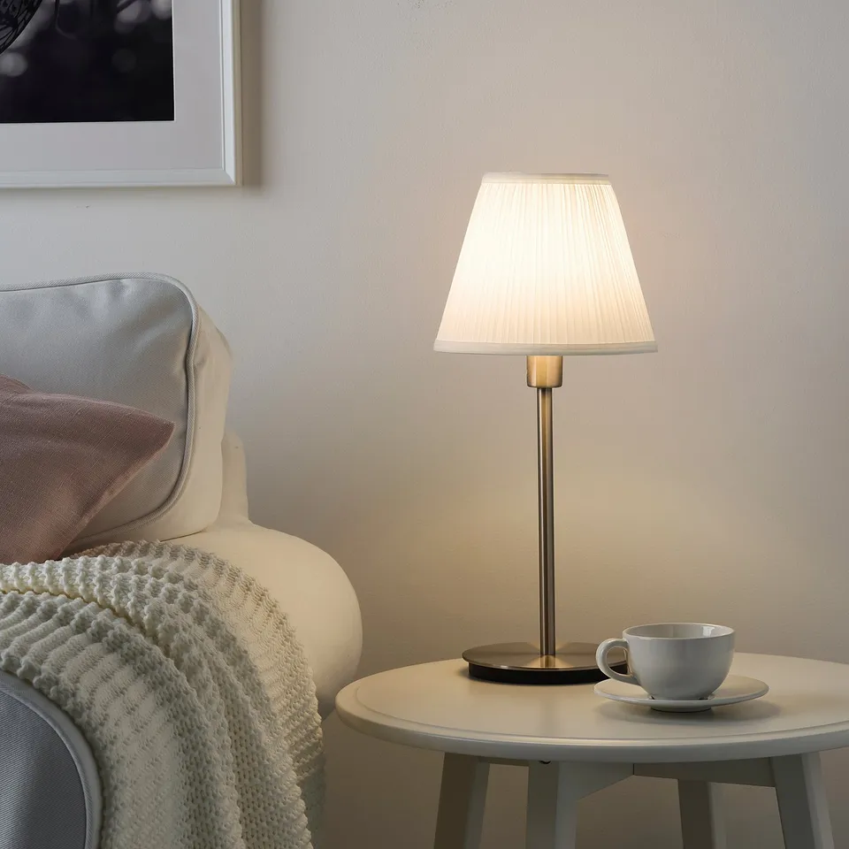 Cosy corner with SKAFTET lamp on table. Armchair with knitted blanket, coffee cup. Lamps small, white shade on top.